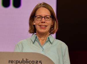 Anne Brorhilker auf der Republica25: Eine l&auml;chelnde Person mit Brille steht vor einem Rednerpult bei einer Veranstaltung, mit buntem Hintergrund.