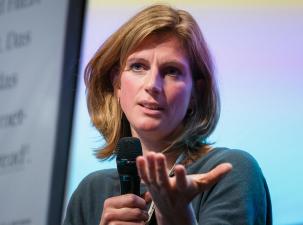 Maja G&ouml;pel auf der re:publica 19 - Frau mit blonden Haaren h&auml;lt ein Mikrofon, sie gestikuliert nachdenklich w&auml;hrend einer Diskussion. Bunte Hintergrundbeleuchtung.