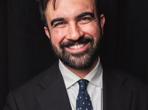 Zohran Mamdani tr&auml;gt einen schwarzen Anzug und ein wei&szlig;es Hemd und l&auml;chelt vor einem schwarzen Hintergrund freundlich in die Kamera.