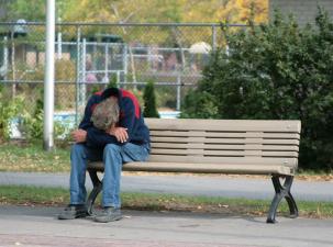 Ein Mann sitzt eingesunken auf einer Bank, den Kopf auf den Armen.