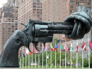 The Knotted Gun by Carl Fredrik Reutersward, UN New York - Eine gro&szlig;e, verdrehte Waffe aus Bronze steht in einer Stadtlandschaft mit Flaggen im Hintergrund.