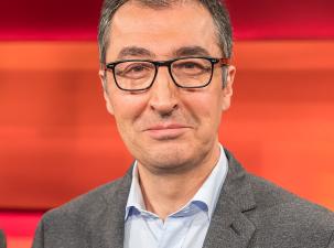 Cem &Ouml;zdemir in 2020 - Ein Mann mit Brille und einem grau-melierten Anzug, l&auml;chelt vor einem orangefarbenen Hintergrund.