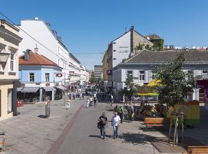 Belebte Stra&szlig;e mit Gesch&auml;ften, Caf&eacute;s und Spazierg&auml;ngern unter klarem blauen Himmel.