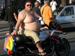 Ein b&auml;rtiger Mann sitzt ohne Oberteil auf einem Motorrad, tr&auml;gt Sonnenbrillen und hat eine Regenbogenflagge dabei.