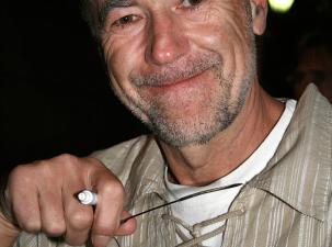 Der &ouml;sterreichischen Musiker Wilhelm 'Willi' Resetarits (Wien 2007) - Ein l&auml;chelnder Mann h&auml;lt eine Brille in der Hand. Er tr&auml;gt ein lockeres, gestreiftes Hemd und hat einen grauen Bart. Hintergrund ist unscharf.