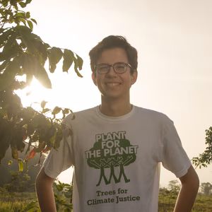 Junger Mann mit Brille steht in einem Garten, tr&auml;gt ein T-Shirt mit dem Aufdruck &bdquo;Plant for the Planet&ldquo; und l&auml;chelt.