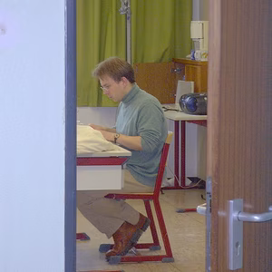 Eine Person sitzt an einem Tisch, n&auml;ht und konzentriert sich, w&auml;hrend im Hintergrund ein gr&uuml;ner Vorhang und eine N&auml;hmaschine sichtbar sind.