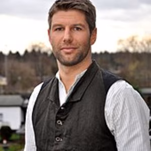 Ein junger Mann mit braunen Haaren, in traditioneller Kleidung, steht vor einer unscharfen Landschaft unter bew&ouml;lkten Himmel.