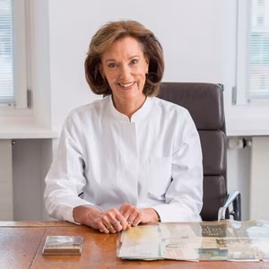 Eine l&auml;chelnde Frau in einem wei&szlig;en Kittel sitzt an einem Tisch mit Dokumenten, in einem modernen, hellen B&uuml;ro.