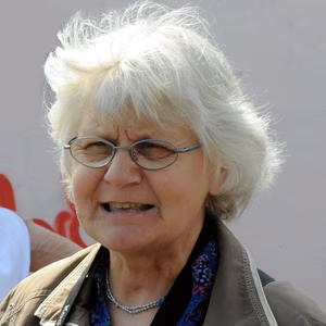 Eine &auml;ltere Frau mit grauem, lockigem Haar und Brille, die mit einem Ausdruck von Besorgnis l&auml;chelt.