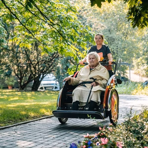 Eine &auml;ltere Frau sitzt in einem Rollstuhl, der von einer Betreuerin auf einem gepflasterten Weg in einem Garten geschoben wird.