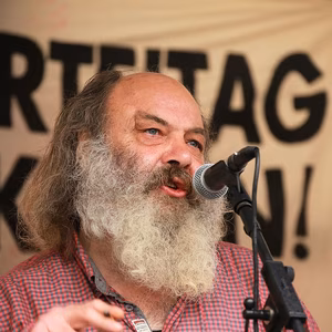 Ein Mann mit grauem Bart spricht leidenschaftlich in ein Mikrofon, w&auml;hrend ein Banner im Hintergrund h&auml;ngt.