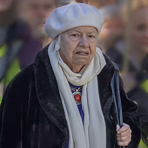 Eine &auml;ltere Frau mit einem wei&szlig;en Hut und einem schwarzen Mantel, umgeben von Menschen in auff&auml;lliger gelber Kleidung.