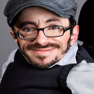 Ein l&auml;chelnder Mann mit Brille und grauem Hut, tr&auml;gt einen schwarzen Pullover &uuml;ber einem wei&szlig;en Hemd, vor neutralem Hintergrund.