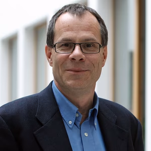 Ein l&auml;chelnder Mann mit Brille und zur&uuml;ckgek&auml;mmtem Haar tr&auml;gt einen Anzug in einem modernen Innenraum.