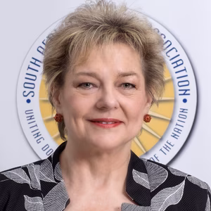 Eine l&auml;chelnde Frau mit blonden Haaren und roten Ohrringen, tr&auml;gt einen grauen Blazer mit Muster, vor einem runden Logo im Hintergrund.