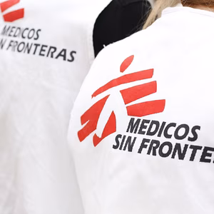 Wei&szlig;e Jacken mit dem Logo von &bdquo;&Auml;rzte ohne Grenzen&ldquo; in Rot und Schwarz, getragen von zwei Personen im Hintergrund.