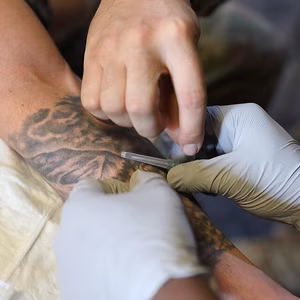 Eine Hand voller Tattoos erh&auml;lt eine Bearbeitung, w&auml;hrend eine andere Hand mit einer Nadel vorsichtig arbeitet.