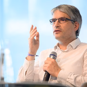 Ein Mann mit Brille und grauen Haaren spricht aktiv in ein Mikrofon, w&auml;hrend er mit der Hand gestikuliert. Heller, moderner Hintergrund.
