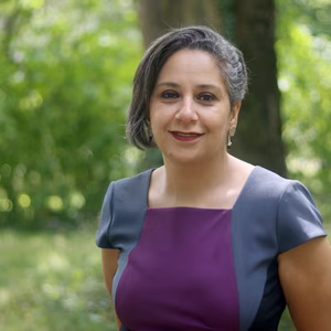 Eine Frau mit kurzen, grauen Haaren steht in einem gr&uuml;nen Park. Sie tr&auml;gt ein lila und graues Kleid und l&auml;chelt freundlich.