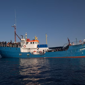 Ein blaues Schiff mit mehreren Personen an Deck, das auf ruhigem Wasser unter klarem Himmel f&auml;hrt.