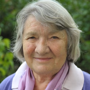 Eine &auml;ltere Frau mit grauen Haaren und einem warmen L&auml;cheln, tr&auml;gt einen lila Schal und steht vor gr&uuml;nem Hintergrund.
