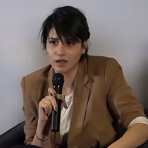 Eine Person mit dunklem Haar und einem blauen Blazer sitzt auf einem Sessel und h&auml;lt ein Mikrofon, w&auml;hrend sie aufmerksam spricht.