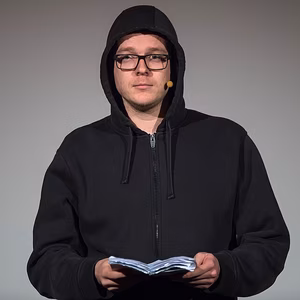 Ein Mann in einem schwarzen Kapuzenpulli h&auml;lt ein Buch in den H&auml;nden und schaut direkt in die Kamera. Hintergrund neutral.