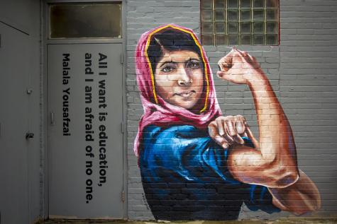 Ein gemaltes Wandbild von Malala Yousafzai auf einer Backsteinmauer. Sie tr&auml;gt ein Kopftuch und schwingt ihren Bizeps, um ihre St&auml;rke zu demonstrieren. Auf einer T&uuml;r daneben sind die Worte "Alles was ich will, ist Frieden" auf Englisch gemalt.