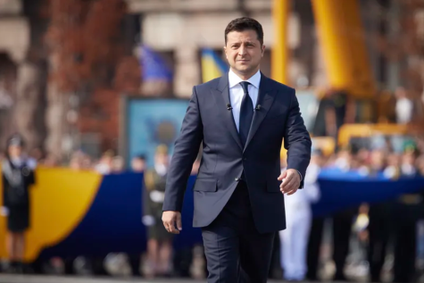 Wolodymyr Selenskyj, der Präsident der Ukraine geht in einem blau-schwarzen Anzug durch eine Truppenparade. Er schaut ernst und weit weg von der Kamera, alles im Hintergrund ist unscharf.