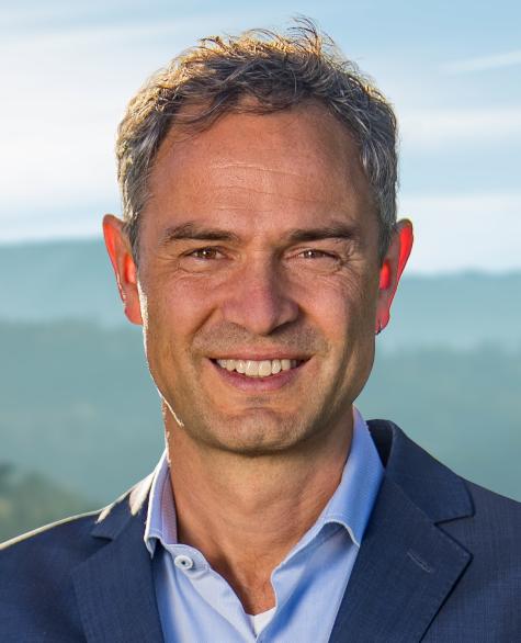 Daniele Ganser - Ein l&auml;chelnder Mann tr&auml;gt einen dunkelblauen Anzug und steht vor einer unscharfen, nat&uuml;rlichen Landschaft.