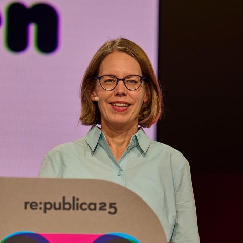 Anne Brorhilker auf der Republica25: Eine lächelnde Person mit Brille steht vor einem Rednerpult bei einer Veranstaltung, mit buntem Hintergrund.