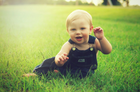 Ein l&auml;chelndes Kleinkind sitzt auf gr&uuml;nem Gras und hebt eine Hand, um zu winken. Die Sonne scheint sanft auf die Szene.