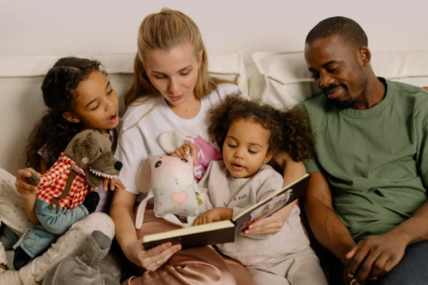 Eine Familie sitzt auf dem Sofa und liest einem kleinen Kind ein Buch vor, umgeben von Stofftieren.