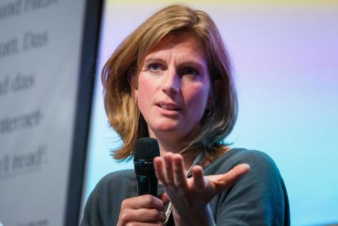 Maja G&ouml;pel auf der re:publica 19 - Frau mit blonden Haaren h&auml;lt ein Mikrofon, sie gestikuliert nachdenklich w&auml;hrend einer Diskussion. Bunte Hintergrundbeleuchtung.