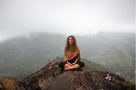 Eine Person mit lockigem Haar sitzt im Schne crossed-leg auf einem Felsen, umgeben von Nebel und einer bergigen Landschaft.