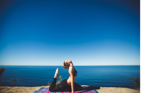 Eine Frau in Yogahaltung sitzt auf einer Matte am Rand einer Klippe mit Blick auf das ruhige Meer und den blauen Himmel.