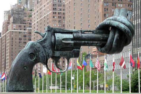 The Knotted Gun by Carl Fredrik Reutersward, UN New York - Eine gro&szlig;e, verdrehte Waffe aus Bronze steht in einer Stadtlandschaft mit Flaggen im Hintergrund.