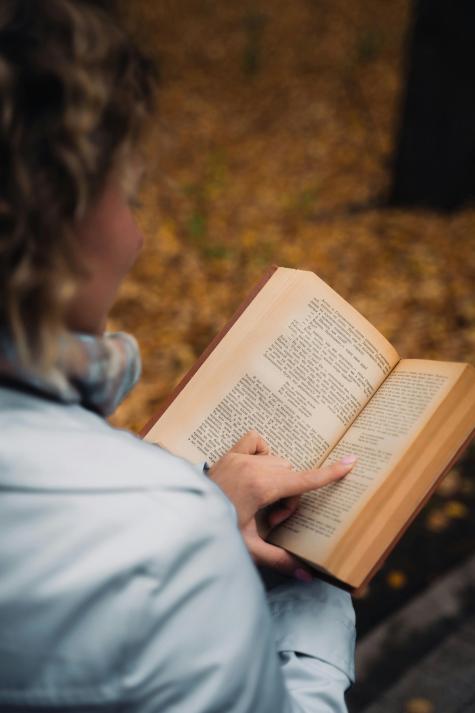 Eine Person mit lockigem Haar h&auml;lt auf einer Bank in einem Park ein aufgeschlagenes Buch in der Hand, umgeben von fallenden Bl&auml;ttern.