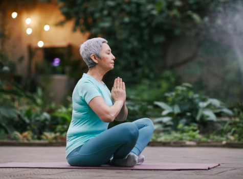 Eine &auml;ltere Frau mit grauem Haar sitzt in einer Meditationspose auf einer Yogamatte, umgeben von gr&uuml;nen Pflanzen.