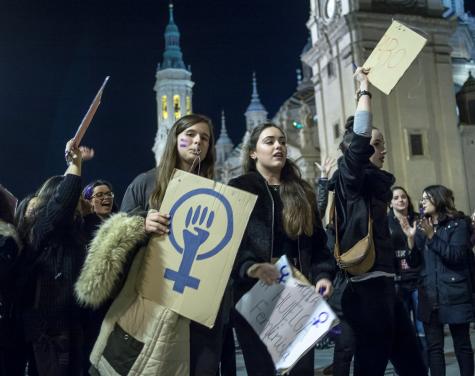 Demonstrierende Frauen mit Schildern, darunter ein feministisches Symbol, vor einer monumentalen Architektur bei Nacht.