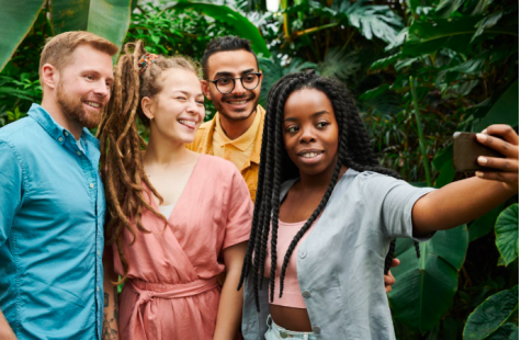 Vier Freunde l&auml;cheln und posieren gemeinsam f&uuml;r ein Selfie vor einer &uuml;ppigen, gr&uuml;nen Pflanzenkulisse.