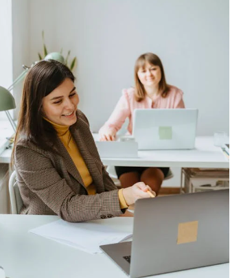 Zwei Frauen arbeiten in einem modernen B&uuml;ro. Eine Frau l&auml;chelt w&auml;hrend sie vor einem Laptop sitzt, die andere im Hintergrund.