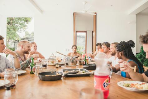 Eine Gruppe von Menschen sitzt an einem langen Tisch beim Essen und Unterhalten. Es sind verschiedene Gerichte und Getr&auml;nke sichtbar.