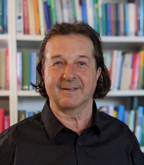 Hans Holzinger - Mann mit braunen Haaren und einem leichten L&auml;cheln, steht vor einem B&uuml;cherregal mit bunten B&uuml;chern.