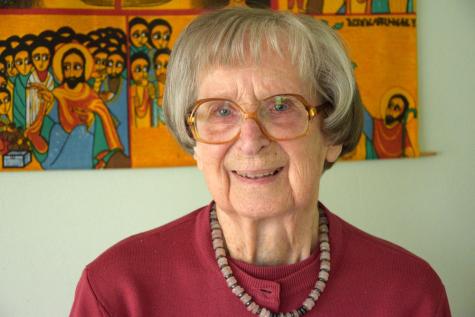 Hildegard Goss-Mayr - Eine &auml;ltere Frau mit Brille und kurzen grauen Haaren l&auml;chelt vor einem farbenfrohen Wandbild. Sie tr&auml;gt einen roten Pullover.