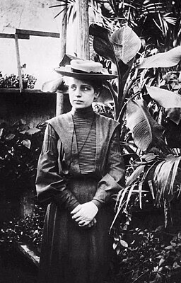 Eine Frau in einem langen, dunklen Kleid und einem Hut steht zwischen Pflanzen in einem gr&uuml;nen Garten.