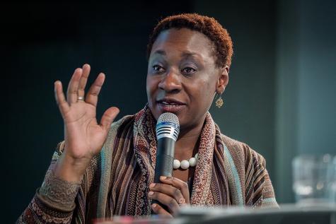 Eine Frau mit kurzen, lockigen Haaren spricht am Mikrofon, hebt eine Hand und tr&auml;gt ein farbenfrohes Oberteil.