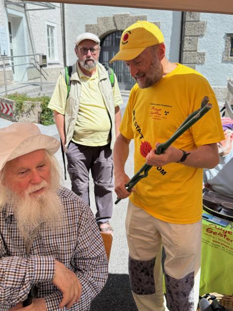 Matthias Reichl, Euratom, Wackersdorf Denkmal in Salzburg - Ein Mann mit einem langen wei&szlig;en Bart sitzt, w&auml;hrend ein anderer Mann mit gelbem T-Shirt und einer Angelrute spricht. Im Hintergrund steht ein dritter Mann.