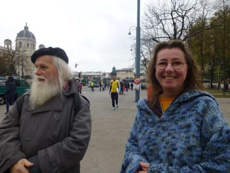Rosi Krenn mit Matthias Reichl in Wien 2014 - Ein &auml;lterer Mann mit langem wei&szlig;en Bart und eine Frau mit lockigem Haar in einem blauen Pullover stehen in einem Stadtpark, umgeben von Menschen.